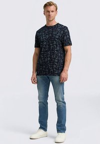 Marinblå t-shirt med abstrakta ljusblå mönster, korta ärmar och avslappnad passform, kombinerad med ljusblå jeans och vita sneakers.