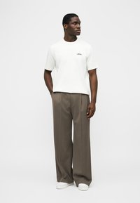 Άνδρας με λευκό κοντομάνικο πουκάμισο, καφέ παντελόνι με φαρδιά μπάρα, λευκά αθλητικά παπούτσια, στέκεται με τα χέρια στις τσέπες, κοιτάζοντας προς τα δεξιά σε απλό φόντο.