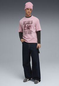 Tricou roz cu imprimeu negru, purtat peste un top cu mâneci lungi închise la culoare. Jeans deblocați largi de culoare închisă și adidași gri. Căciulă roz.