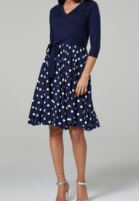 Mörkblå klänning med prickar, med omslagstopp och trekvartsärmar, åtdragen i midjan med ett matchande bälte. Utsvängd kjoldesign.