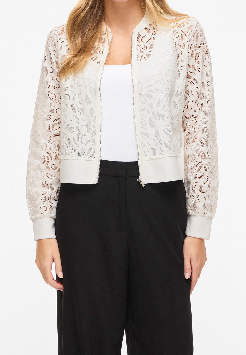 Femme portant une veste bomber en dentelle blanche sur un haut blanc, associée à un pantalon noir taille haute à jambes larges, debout devant un fond blanc.