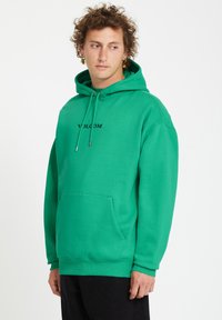 Groene hoodie van katoen, met een voorzak, een trekkoordkap en een zwart logo "VOLCOM" op de borst. Zachte textuur.