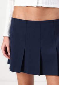 Gonna plissettata blu navy realizzata in tessuto morbido, con pieghe nette e uniformemente distanziate e un orlo pulito. Abbinata a una maglietta bianca a coste.