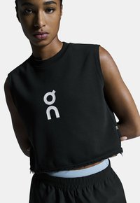 Schwarzes, kurz geschnittenes Tanktop mit ärmellosem Design, mit einem weißen Logo auf der Vorderseite. Roher Saum und strukturierter Stoff. Kombiniert mit schwarzen Shorts.