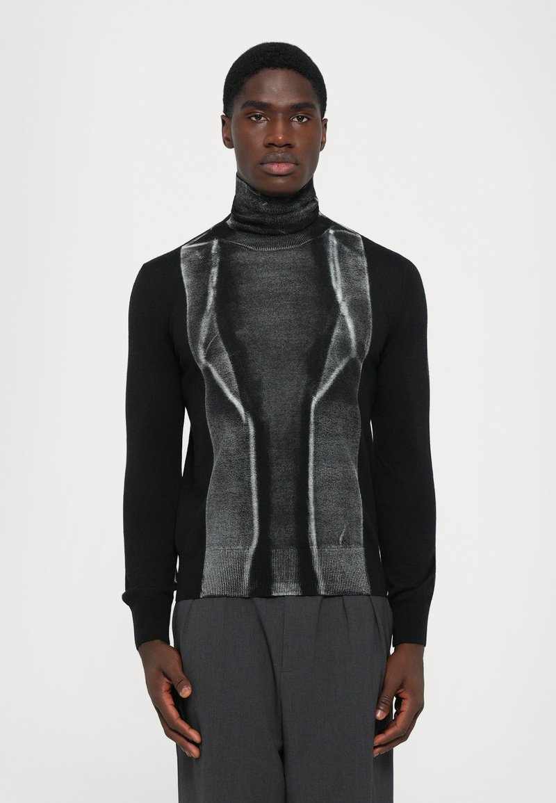 Giovane uomo che indossa un maglione nero a collo alto con un motivo geometrico argentato lucido sul davanti, abbinato a pantaloni grigio scuro.