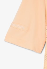 Tissu pêche à manches courtes avec une pièce rectangulaire cousue portant l'inscription "LACOSTE" près de l'ourlet de la manche.