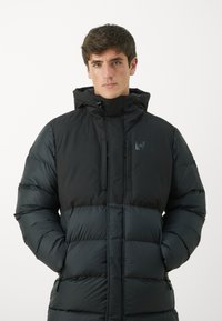 Jachetă puffer neagră cu guler înalt și glugă ajustabilă. Prezintă buzunare cu fermoar și un amestec de texturi mate și lucioase.