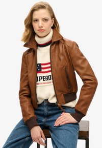 Giacca bomber in pelle marrone con chiusura a zip, polsini a coste e colletto, abbinata a un maglione in maglia crema con grafiche rosse e blu.