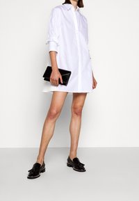 Abito camicia bianco con maniche arrotolate, abbinato a una pochette nera e scarpe nere con borchie argentate. Tessuto liscio.