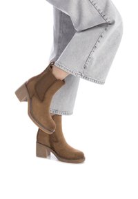 Stivaletti alla caviglia in suede marrone con tacco a blocco, pannelli elastici laterali e cuciture a contrasto, indossati con jeans a gamba larga grigi chiari.