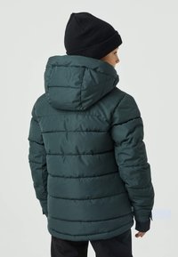 Ciemnozielona kurtka puchowa z kapturem, pikowanym wzorem i elastycznymi mankietami. Tył ma gładką fakturę i subtelny akcent marki.