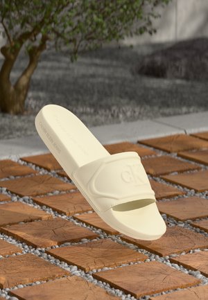 Sandale na klizanje svijetle boje Calvin Klein s utisnutim "ck" logotipom, prikazane iznad drvenih i šljunčanih vanjskih pločica s mutnom pozadinom drveća.