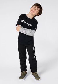 Χαμογελαστό παιδί που στέκεται με τα χέρια σταυρωμένα, φοράει μαύρη μπλούζα Champion, μαύρο παντελόνι φόρμας με λευκό λογότυπο και ελιόχρωμα αθλητικά παπούτσια.