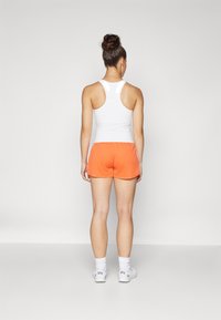 Weißes sportliches Tanktop und orangefarbene Shorts, mit einer figurschmeichelnden Passform und glatter Textur. Weiße Schuhe mit Stoffsocken, von hinten betrachtet.