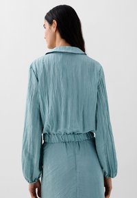 Lysblå, krøllet stofjakke med krave, elastisk talje og lange ærmer. Glat, flydende tekstur med minimal detaljering.