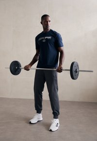 Um homem em uma t-shirt azul marinho, calças de moletom cinza e tênis brancos segura uma barra de peso preta, contra um fundo de parede de cor neutra.
