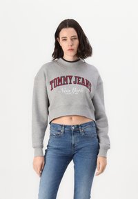 Felpa grigia corta con scritte rosse "TOMMY JEANS" e "New York", colletto a costine. Abbinata a jeans skinny blu.