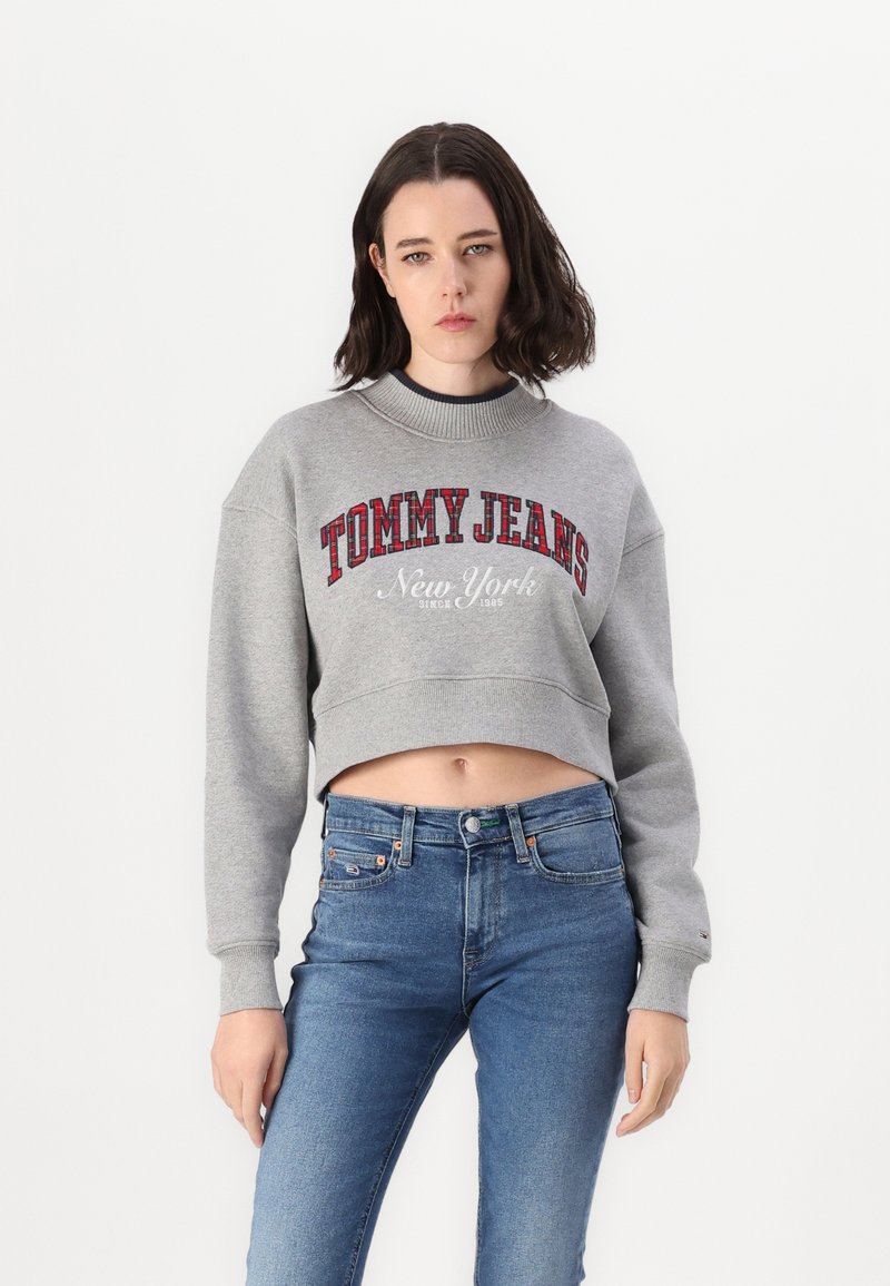 Felpa grigia corta con scritte rosse "TOMMY JEANS" e "New York", colletto a costine. Abbinata a jeans skinny blu.
