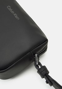 Bolsa de cuero negro con una textura suave, que presenta un cierre metálico y el logo en relieve de "Calvin Klein" en la parte frontal.