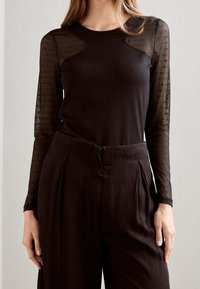 Frau trägt ein schwarzes, figurbetontes Oberteil mit transparenten, gepunkteten langen Ärmeln und hochtaillierten, strukturierten schwarzen Hosen vor einem neutralen Hintergrund.