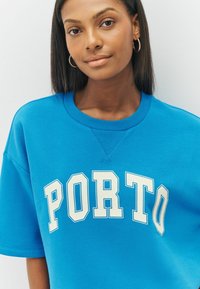 Jauna moteris, dėvinti auksinius auskarus ir ryškiai mėlyną marškinėlį su dideliais baltais žodžiais "PORTO".