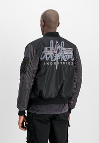 Mustmust must, mille eest, hall tikkidega "alpha INDUSTRIES" logo, soonikuvarrukad ja -äär, sile nailonmaterjal ja utiliittasku detail.
