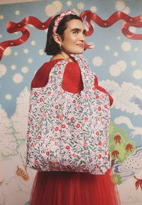 Virágos szövet bevásárlótáska rózsaszín háttérrel, piros virágokkal és zöld levelekkel díszítve. Puha anyagból készült, lekerekített fogantyúkkal, párosítva egy piros kötött pulóverrel.