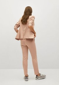 Blazer rose poudré sur mesure avec un détail de pli, associé à un pantalon slim assorti et des chaussures de sport grises, vu de dos.