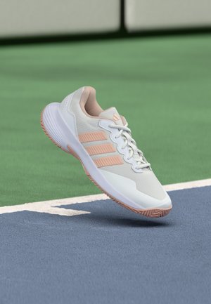 Scarpa sportiva Adidas bianca e rosa chiaro sospesa sopra la superficie di un campo da tennis blu e verde vicino alla linea di confine bianca.