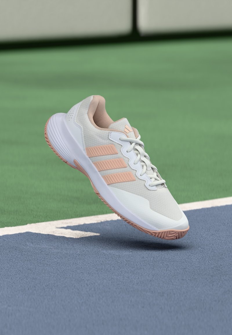 Scarpa sportiva Adidas bianca e rosa chiaro sospesa sopra la superficie di un campo da tennis blu e verde vicino alla linea di confine bianca.