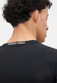 Cămașă sport Nike Pro neagră cu guler rotund, cu cusături ale logo-ului alb pe spate și material neted, elastic.