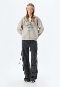 Sudadera con cremallera de color gris claro con un estampado gráfico y cordones, combinada con pantalones cargo negros holgados que tienen bolsillos con solapa y lazos.