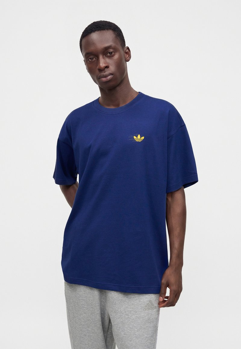 Hombre con una camiseta azul con un pequeño logo amarillo y pantalones deportivos Adidas gris claro, de pie sobre un fondo blanco liso.