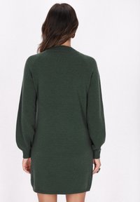 Strickkleid in Dunkelgrün mit langen, schulterfängigen Ärmeln und rundem Ausschnitt. Glatte Textur, mittlere Oberschenkellänge und seitliche Schlitze.