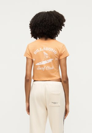 Άτομο με σγουρά μαλλιά φοράει πορτοκαλί crop top με λευκό "Billabong Surf Club" και γραφικό με σερφ, συνδυασμένο με κρεμ παντελόνι με πίσω τσέπη.