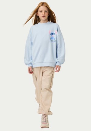 Felpa blu chiaro con una grafica di un personaggio in rosa e blu. Abbinata a pantaloni cargo beige e sneakers con un design testurizzato.