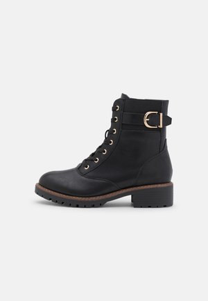 Bottine noire en cuir avec lacets sur le devant, talon épais, bordure de semelle marron et bride à boucle dorée sur le côté extérieur.
