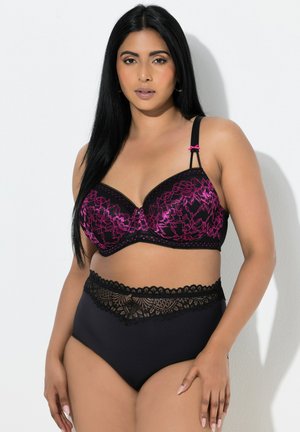 Kvinde med langt sort hår iført sort og lilla blomstret blonde-bh og sorte trusser med høj talje og blonda detaljer, stående foran hvid baggrund.