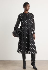 Abito nero con pois bianchi, maniche lunghe trasparenti, scollo rotondo, vita cinchata e cintura. Abbinato a stivali neri con tacco al ginocchio e una pochette nera.