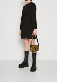 Musta pitkähiha dressi, jossa on pitsikoristeita, vyötärö on kapeampi, ja keltainen leopardikuosiset käsilaukku. Mallilla on jalassaan polven ylle ulottuvat mustat platform-kengät.