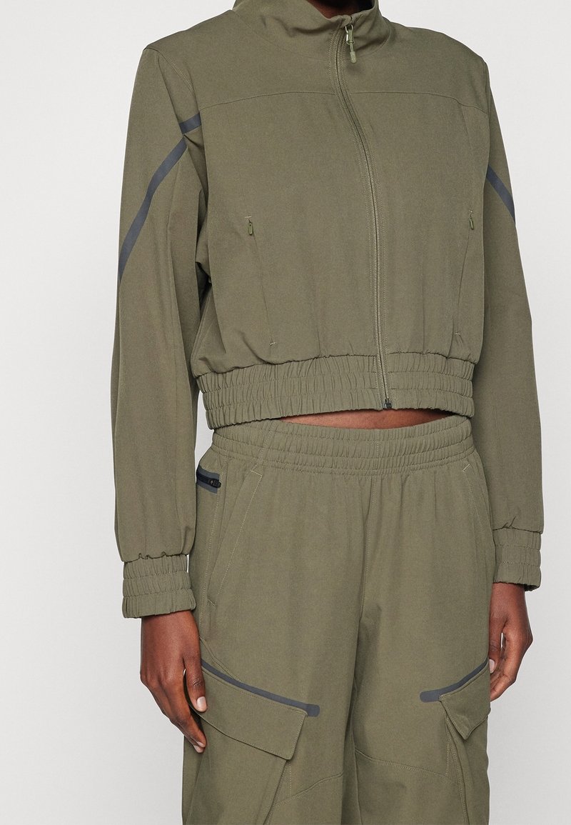 Olive green cropped jacket with zip closure, side pockets, and elastic cuffs. Features gray stripes along the shoulders. Soft, textured fabric.