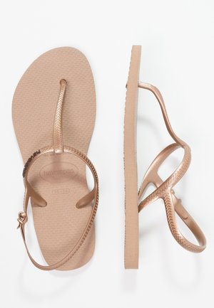Een paar beige platte sandalen met gestructureerde zolen en dunne metalen roségouden bandjes, inclusief een teenlus en een enkelriem met gesp.