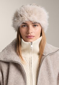 Világos bézs műszőrme sapka bolyhos textúrával; a modell krémszínű cipzáras pulóvert visel bordázott mintával, puha bézs dzseki alatt.
