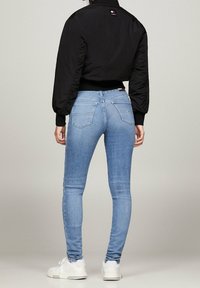 Mujer vestida con jeans ajustados de color azul claro, chaqueta corta negra y zapatillas blancas, de pie de espaldas frente a un fondo sencillo.