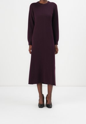 Femme portant une robe midi à manches longues de couleur violet foncé, associée à des chaussures à talons noires, debout devant un fond blanc uni.