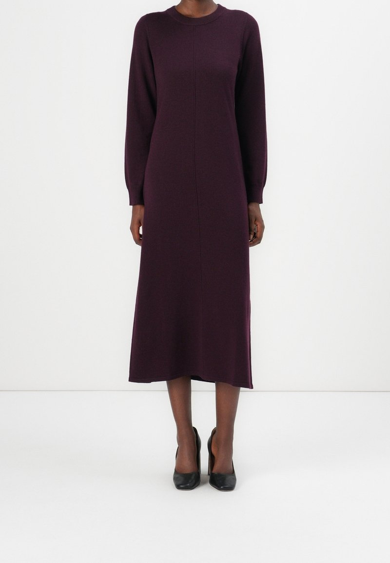 Femme portant une robe midi à manches longues de couleur violet foncé, associée à des chaussures à talons noires, debout devant un fond blanc uni.
