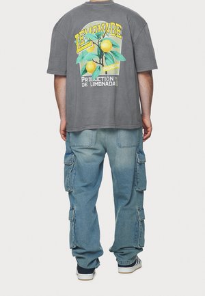 Szara bawełniana koszulka z grafiką cytryny na plecach, w połączeniu z jasno niebieskimi jeansami cargo z bocznymi kieszeniami i luźnym krojem.