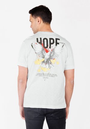 Hombre con camiseta blanca con un gran gráfico de una paloma voladora, salpicaduras de pintura roja y amarilla, y la palabra "ESPERO" en la parte trasera.