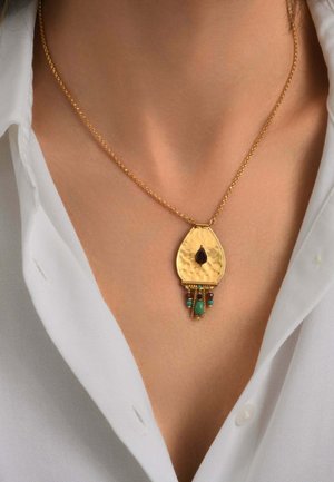 Collier chaîne en or avec un pendentif en or martelé présentant une pierre centrale sombre et de petites perles turquoises et marron suspendues, porté avec une chemise blanche.