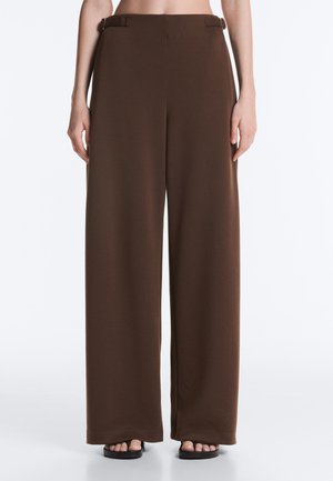 Kobieta ubrana w brązowe, szerokie spodnie z wysokim stanem z bocznymi klamrami, stoi z rękami swobodnie zwisającymi wzdłuż ciała.